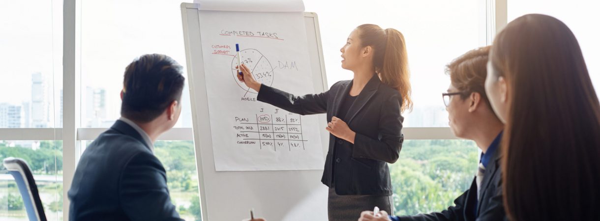 Attractive Asian businesswoman with ponytail pointing at diagram on marker board while holding working meeting in spacious boardroom